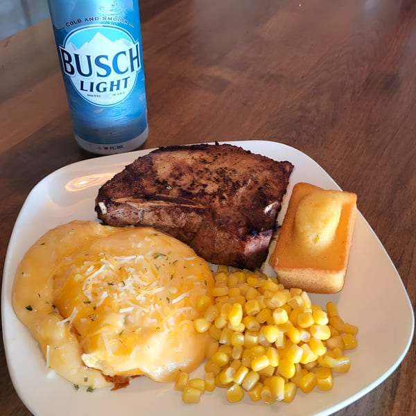 A Lodge on Main meal with a grilled pork chop, cheesy potatoes, corn, cornbread, and a Busch Light.