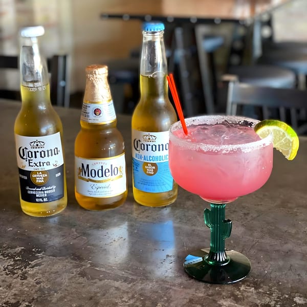 Three beer bottles and a pink margarita in a cactus glass with a lime on a bar top.