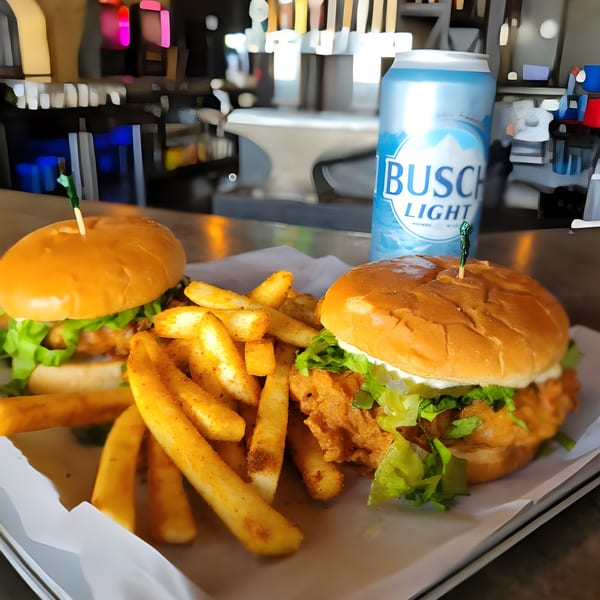 Two crispy chicken sandwiches with lettuce, fries, and a Busch Light beer can on a bar.