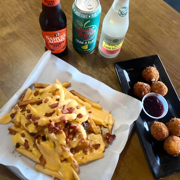 Loaded fries with cheese and bacon, fried balls with dipping sauce, and three bottled drinks on a wooden table.