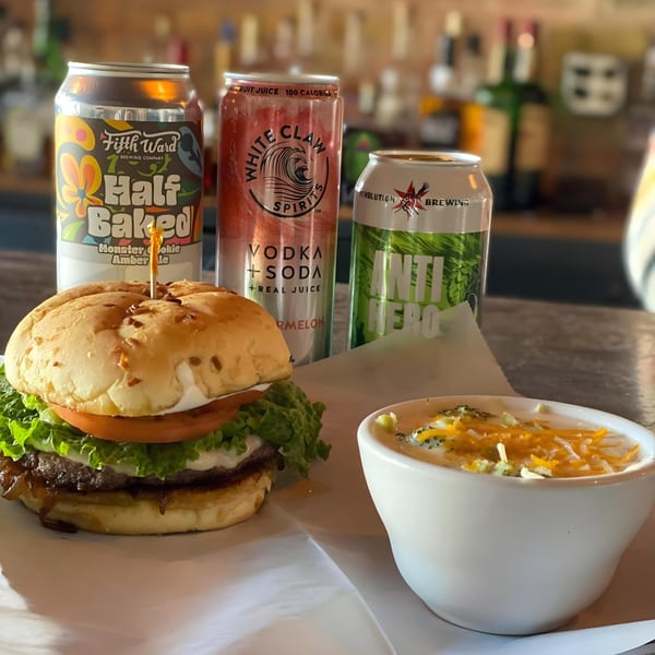 A burger with lettuce, tomato, and caramelized onions, alongside a bowl of broccoli cheddar soup and three canned beverages.