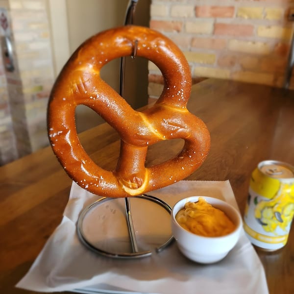 A large, salted pretzel on a stand with a side of cheese dip and a drink can.